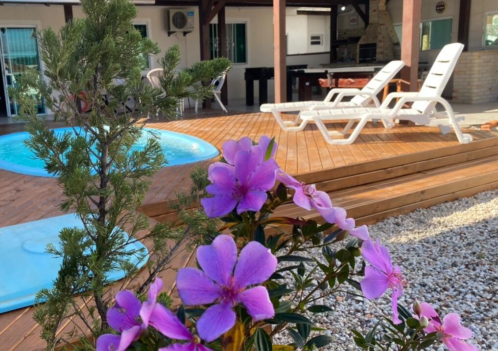 A serene outdoor area features a small pool and sun loungers on a wooden deck. Vibrant purple flowers and a greenery enhance the setting, creating a peaceful environment for relaxation.
