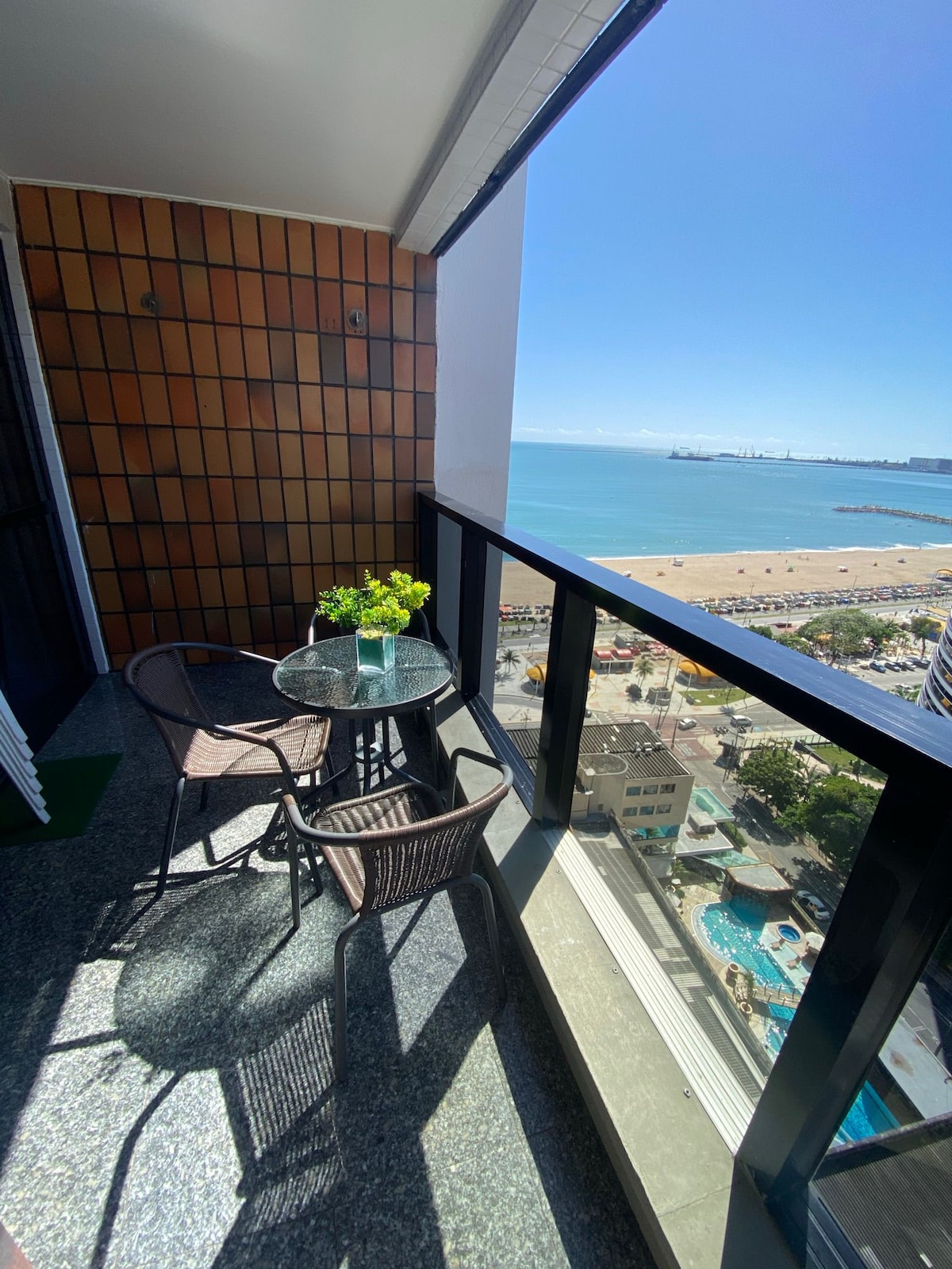 A private balcony features a small glass table with two wicker chairs, surrounded by a scenic view of the ocean. The coastline can be seen along with sandy beaches and clear blue waters under a bright sky.