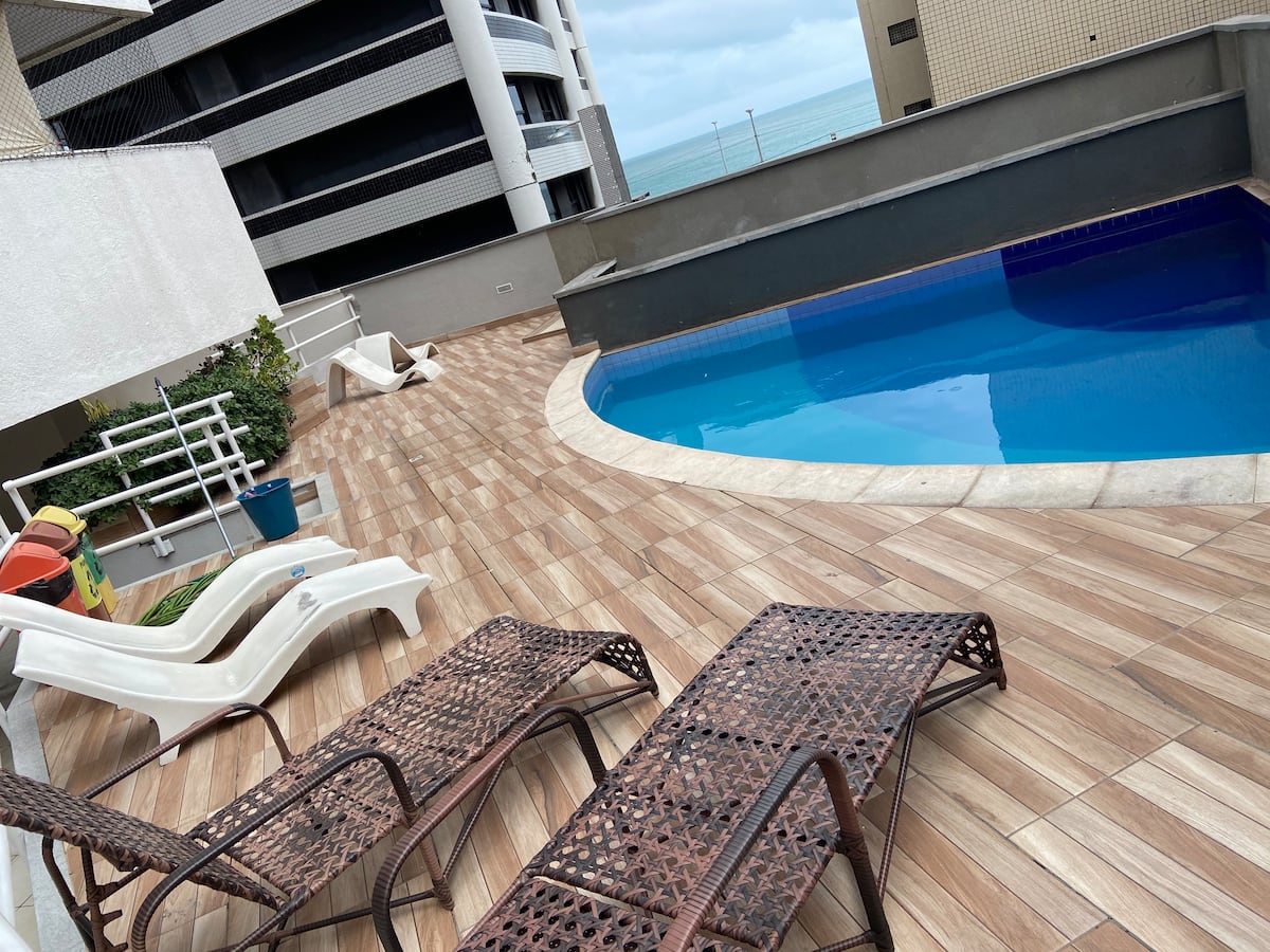 A pool area features a curved swimming pool surrounded by a spacious deck. Two loungers made of wicker are positioned for relaxation. A glimpse of the ocean can be seen in the background, alongside a few plants and outdoor play equipment.