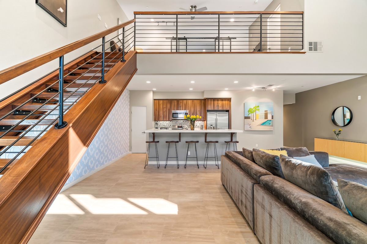 An open-concept living space showcases high ceilings with modern light fixtures and a wooden staircase. The kitchen area is equipped with stainless steel appliances and a bar with seating for three. Natural light fills the room through large windows, creating a bright atmosphere.