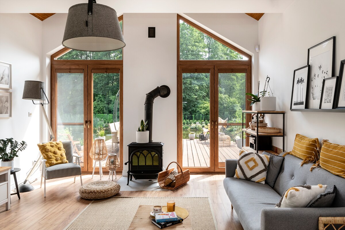 A bright living area features large windows that provide views of the surrounding greenery. A modern gray sofa is paired with a round natural fiber rug. A vintage-style stove and open shelving add character, while light wood accents enhance the spacious atmosphere.