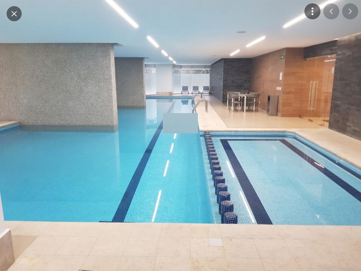 An indoor pool area features a spacious swimming pool with clear blue water, complemented by a separate wading area. Lounge chairs are positioned along the poolside. Natural light floods the space, highlighting the sleek stone walls and modern design elements.