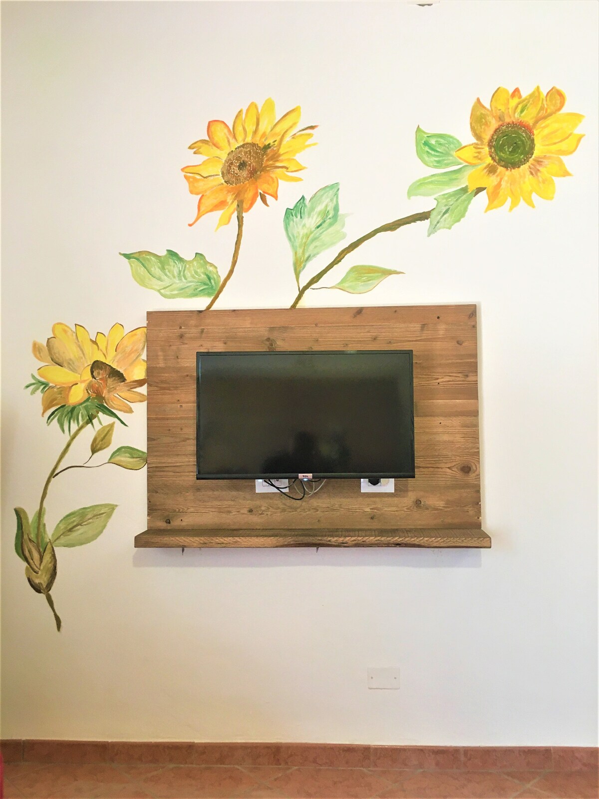A wall-mounted television is framed by a wooden panel, featuring artistic sunflower murals that extend gracefully towards the ceiling. The backdrop is painted in a light color, allowing the vibrant sunflowers to stand out, creating an inviting focal point in the space.