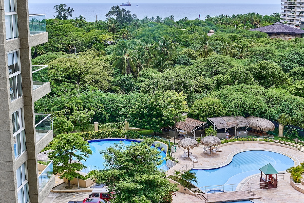 The image captures a scenic view of the lush greenery surrounding the property, with a large swimming pool at the forefront. Several palm trees are visible, and the ocean can be seen in the distance, providing a serene backdrop to the outdoor amenities.