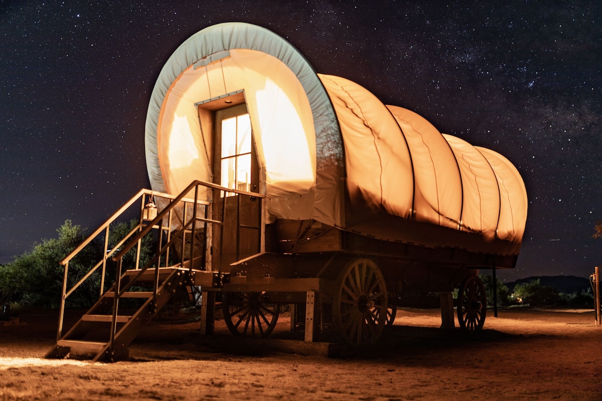 A covered wagon is illuminated by soft light, showcasing its entrance. Steps lead up to the door, enhancing accessibility. The wagon's distinctive rounded top is visible against a night sky filled with stars, creating a tranquil outdoor atmosphere.