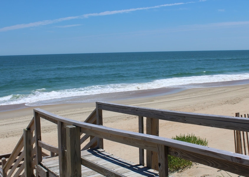 A wooden boardwalk leads towards the sandy beach, framed by low coastal vegetation. The ocean stretches in the background, with gentle waves lapping at the shore under a clear blue sky.