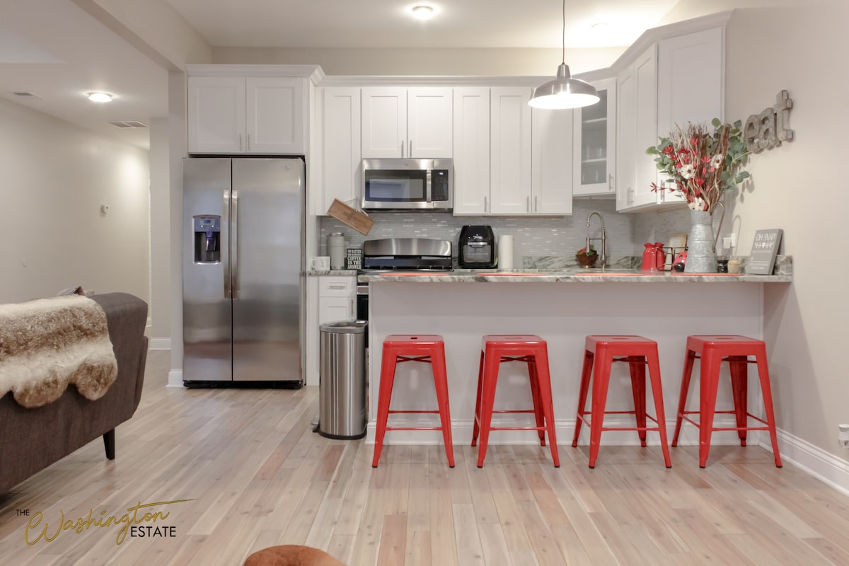 A modern kitchen features white cabinetry and stainless steel appliances, including a refrigerator and microwave. A marble countertop extends to a bar area with three red stools. Decorative elements, including a vase of flowers, enhance the inviting atmosphere.