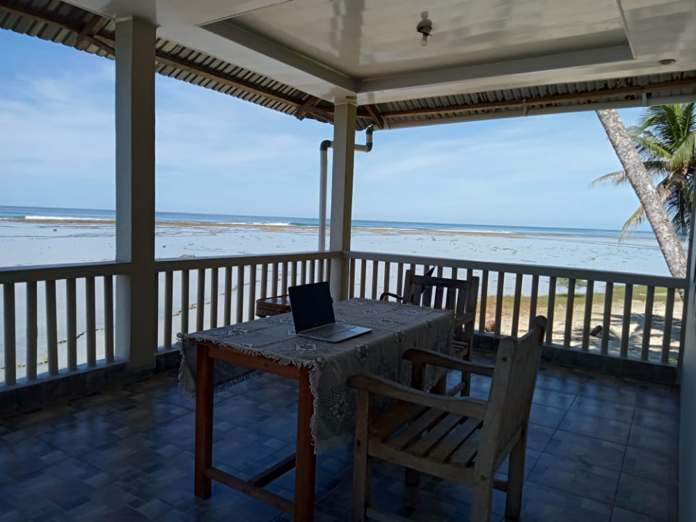 A large terrace showcases a dining table with a lace tablecloth and a laptop, offering expansive views of the beach and ocean. Wooden chairs are positioned around the table, while palm trees can be seen in the background, contributing to the tranquil setting.