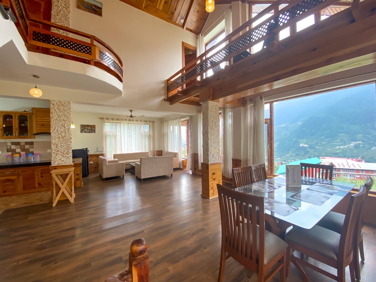 A spacious living area features large windows, providing sweeping views of the surrounding mountains. The wooden furnishings complement the bright interiors, with a dining table set for six. A cozy seating arrangement is positioned nearby, enhancing the welcoming feel of the room.