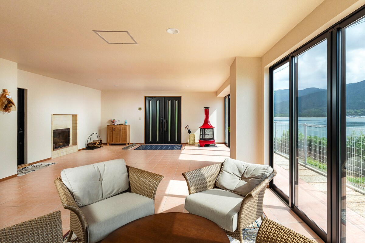 A spacious living area is presented with large windows allowing views of the sea and mountains. Comfortable seating arrangements made of rattan are arranged around a round table. A modern red wood stove is positioned against the wall, enhancing the room's aesthetic.