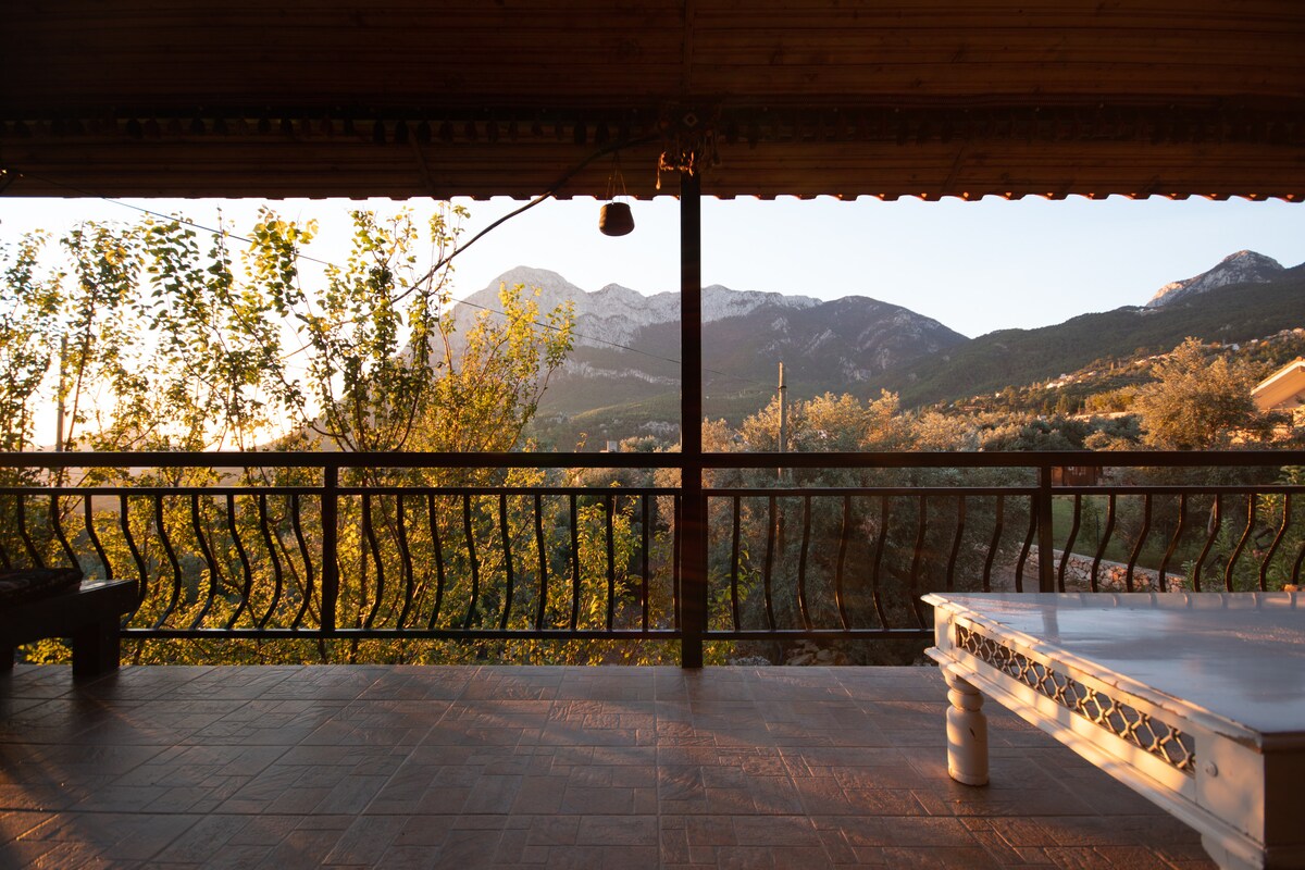 A terrace area features a marble table and wooden seating, overlooking a scenic mountain landscape. Warm sunlight casts a soft glow on the space, highlighting the details of the railing and surrounding greenery.