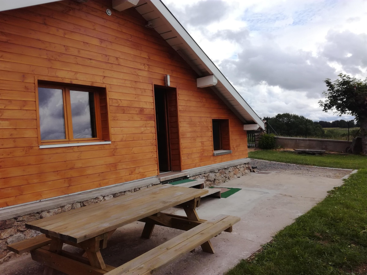 The exterior of the chalet features natural wood siding and a sloped roof. A picnic table is situated on a concrete patio, with green grass surrounding the area. Windows allow for natural light, offering views of the nearby landscape.