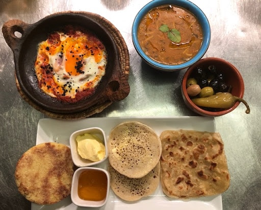 A vibrant food arrangement is presented on a silver surface. A clay dish holds an egg-based dish topped with black seeds, alongside a bowl of curry. Different breads are served with small dishes of butter, honey, and olives, enhancing the variety of the meal.