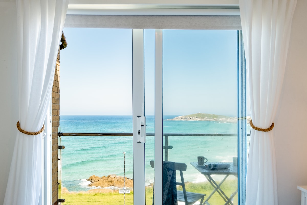 A sliding glass door framed by light, sheer curtains showcases stunning views of Fistral Bay. The tranquil sea stretches toward the horizon, while a small table and chairs are placed on the private terrace, inviting moments of relaxation amidst the coastal scenery.