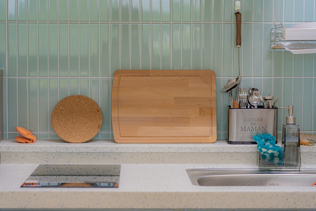 The kitchen area features a light-colored countertop with a built-in cooktop and a stainless-steel sink. A wooden chopping board and a cork mat rest against the backsplash, with various utensils stored in a metal container labeled 'CUISINE DE MAMAM.' A small sponge holder is also visible.