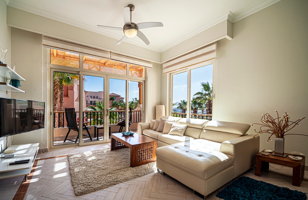 A bright living room features a large sectional sofa and a flat-screen TV. Sliding glass doors open to a balcony with views of palm trees. Natural light fills the space, highlighting the light-colored walls and comfortable atmosphere. A textured rug adds warmth to the room.