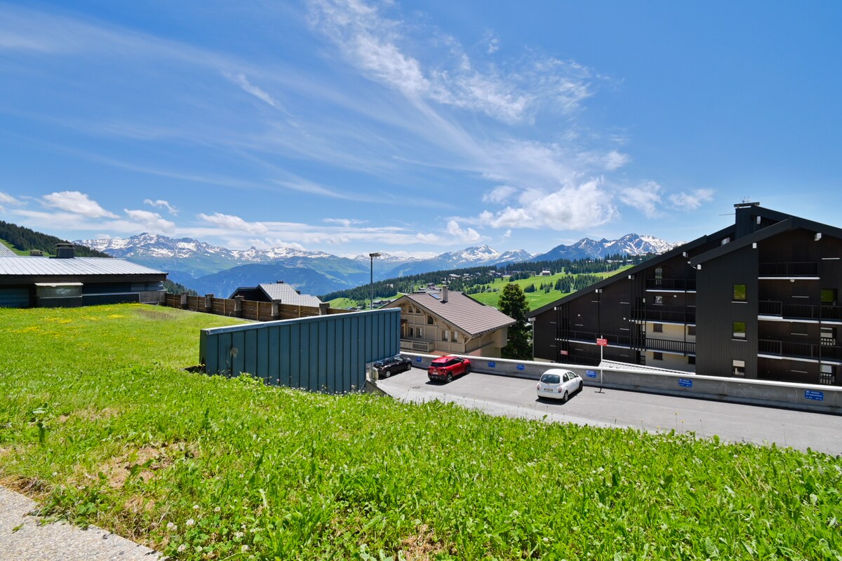A view of the surrounding mountains showcases rolling green fields under a blue sky with scattered clouds. Various buildings, including a multi-story residence and smaller structures, are visible, framed by the inviting landscape. Snow-capped peaks can be seen in the background, accentuating the scenic beauty.