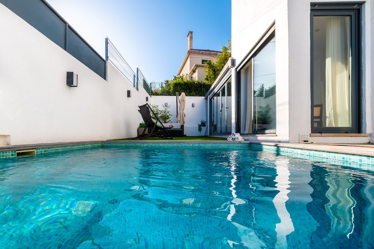 A refreshing swimming pool is depicted, surrounded by a mix of glass walls and white structures. Lounge chairs are visible nearby, under clear blue skies. The pool reflects sunlight, creating a serene ambiance, while greenery adds a touch of nature to the setting.