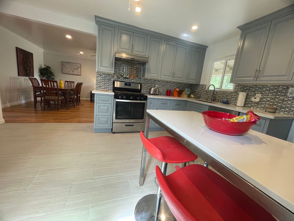 A modern kitchen is furnished with gray cabinetry and a stainless steel stove. A red bowl sits on a kitchen island with red bar stools. A dining area with brown wood furniture is visible in the background, complemented by warm wooden flooring.