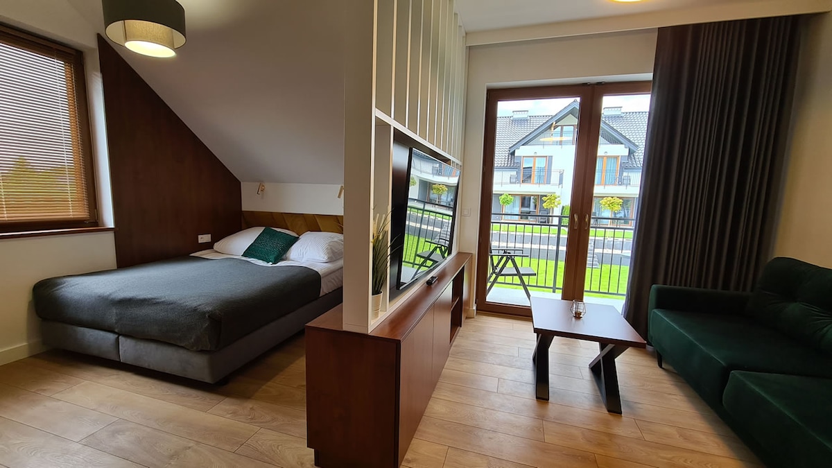 A spacious room is furnished with a large bed and a green sofa. Natural light enters through large sliding doors, which lead to a small balcony overlooking the outdoors. A flat-screen TV is mounted on the wall, and wooden flooring adds a contemporary touch.