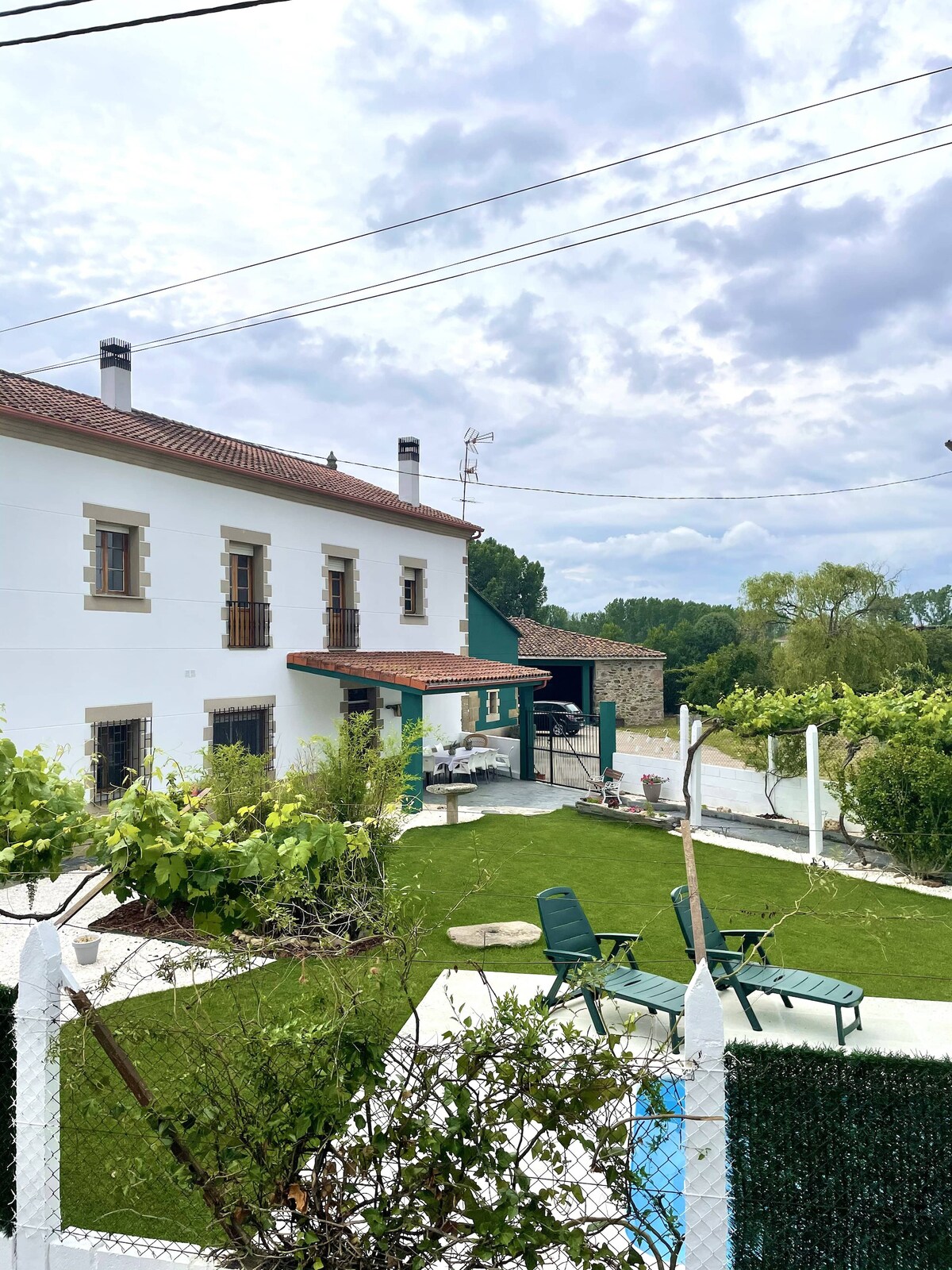 The exterior showcases a spacious garden area with lush greenery and a well-maintained lawn. Several outdoor lounge chairs are positioned for relaxation. A traditional two-story building features a mix of stone and whitewashed walls, with multiple windows that enhance the inviting atmosphere.