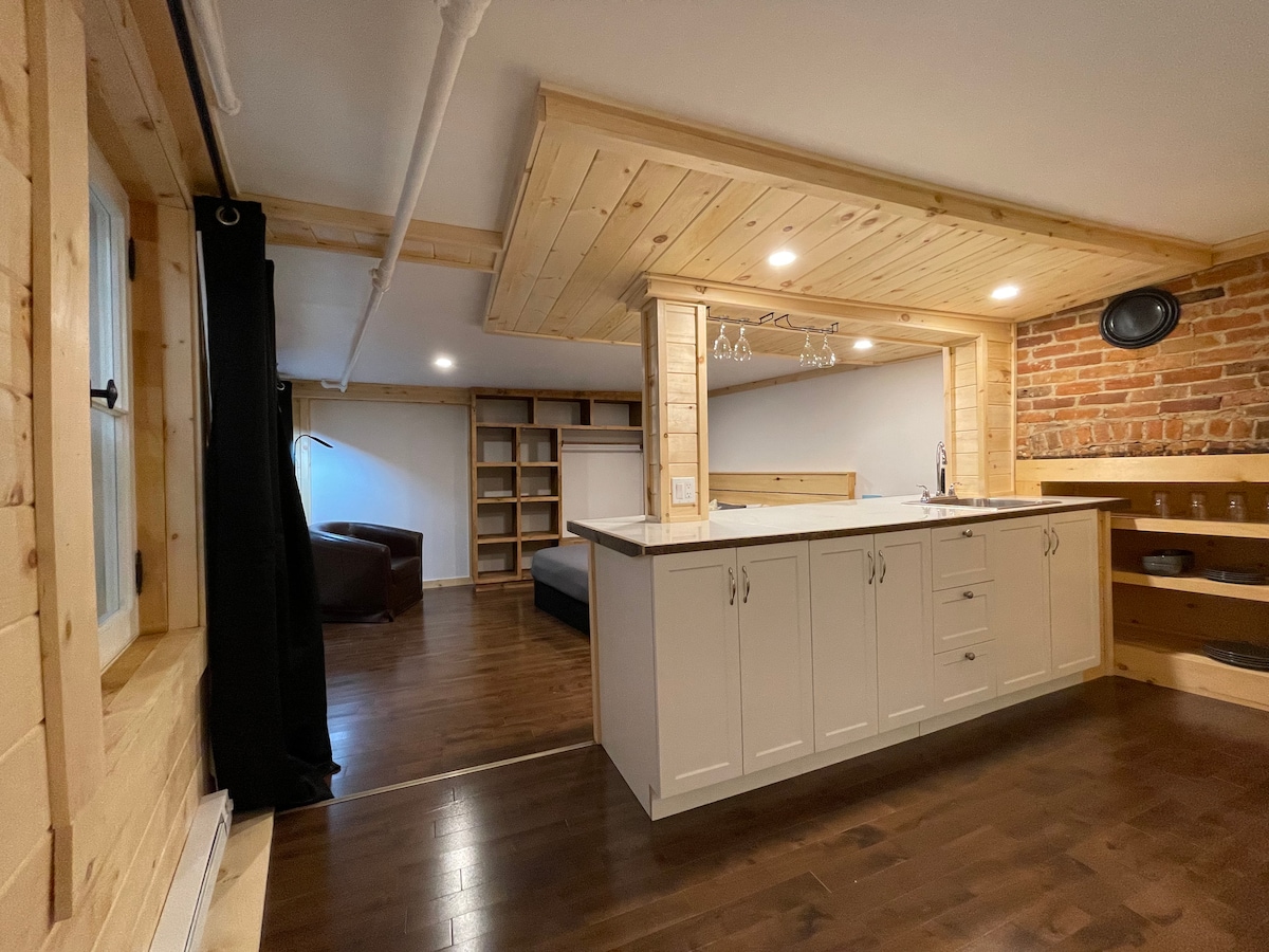 An open loft space features wooden accents throughout, with a modern kitchen area showcasing light cabinetry. A cozy seating area is visible, accompanied by a brick wall that adds texture. Natural wood flooring enhances the room's warmth, while recessed lighting contributes to a well-lit environment.