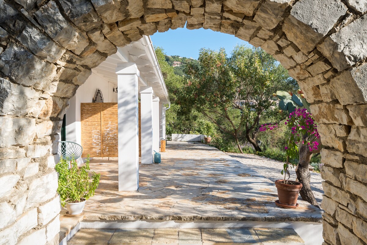 An archway crafted from natural stone frames a spacious terrace area. The floor features a smooth stone surface, and potted plants are positioned around the space. Lush greenery and vibrant flowers create a serene outdoor setting, inviting guests to enjoy the views.