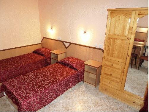 A simple bedroom features two twin beds dressed in deep red bedding. Each bed is flanked by a wooden nightstand. A tall wooden wardrobe with a mirror provides a functional addition, while soft lighting emanates from wall sconces, creating a calm environment.