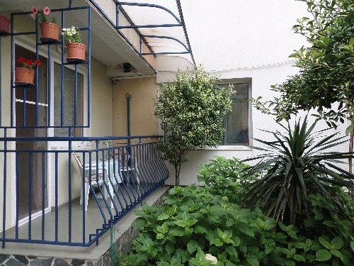 A serene outdoor area features greenery and decorative plants, with a balcony furnished with two chairs and flower pots. A window allows natural light to enter the nearby room, contributing to a calm atmosphere.