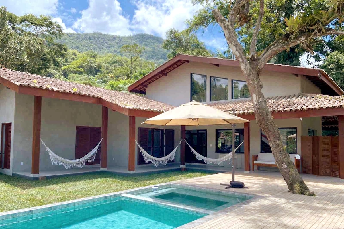 The exterior of the house showcases a spacious deck with a swimming pool, complemented by a large umbrella for shade. Hammocks are seen suspended between trees, surrounded by lush greenery, with mountain views enhancing the tranquil setting.