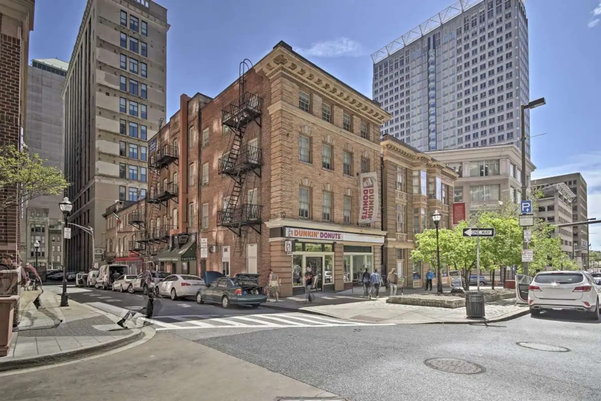 The image displays a historical brick building with fire escapes, located at a city intersection. Nearby modern skyscrapers contrast with the classic architecture. Street signs guide pedestrians, and parked cars occupy the curb lanes, enhancing the urban setting.