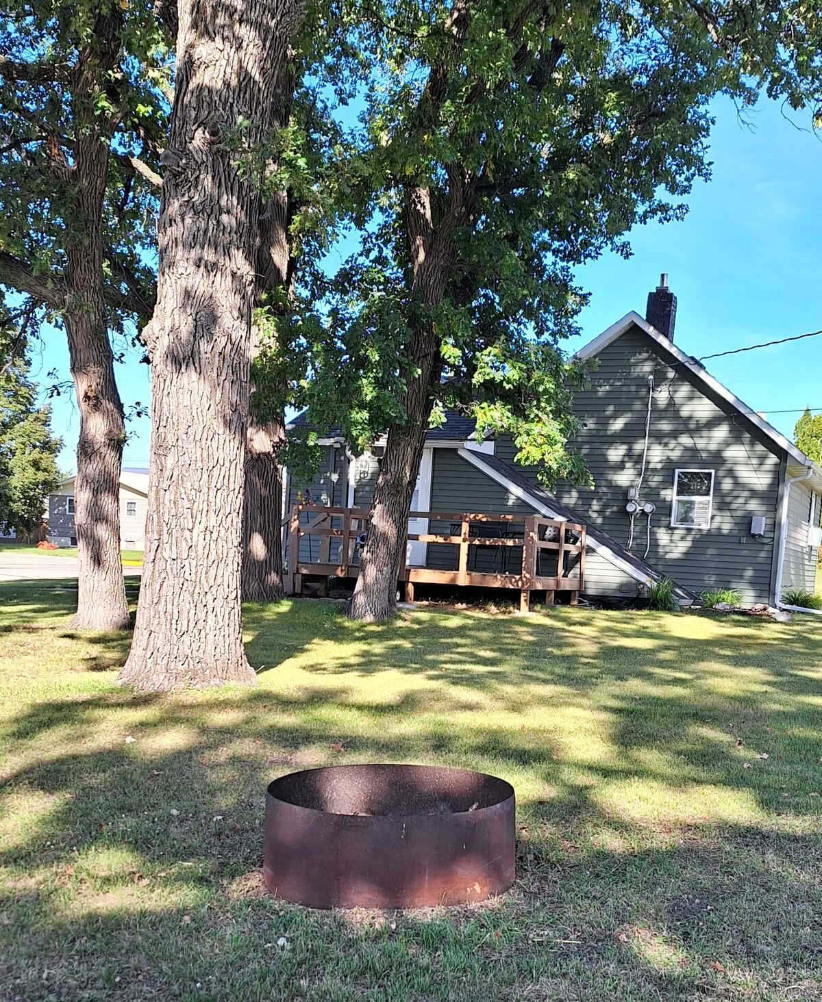 A firepit is nestled in the grassy yard, surrounded by large oak trees. The greenery provides shade, while a well-maintained deck is visible at the back of the vintage home. The exterior displays a muted color scheme with a blend of wood and siding.