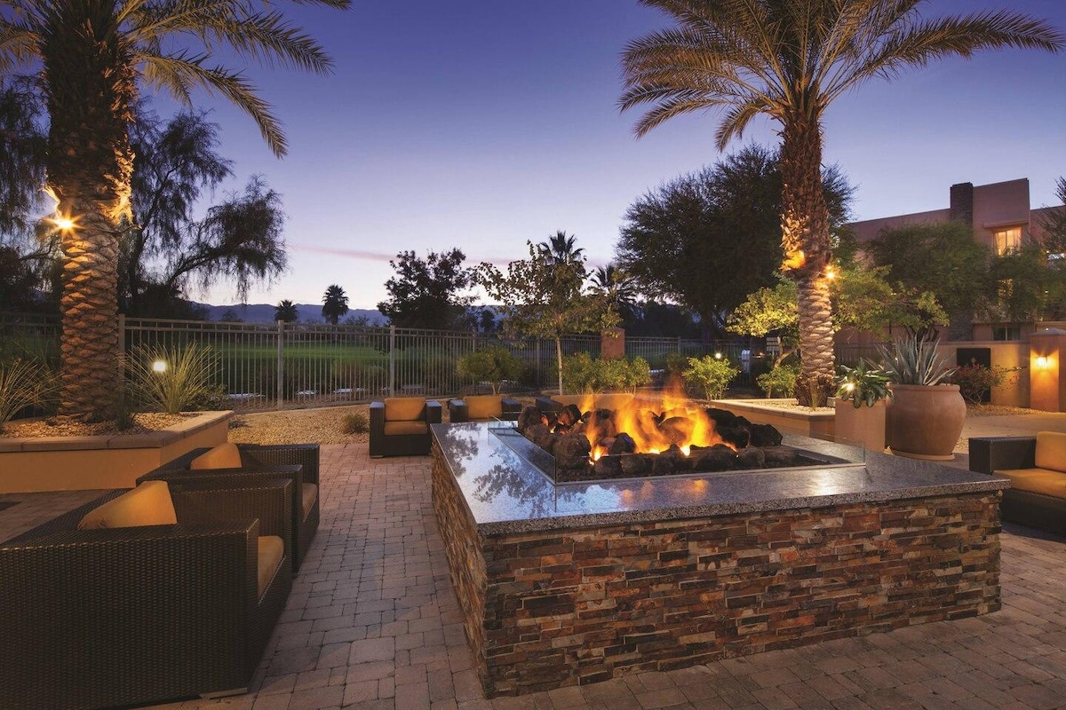An outdoor seating area is illuminated by soft lighting, featuring a stone fire pit surrounded by comfortable seating. Palm trees and landscaped greenery add to the tranquil ambiance. The backdrop includes a view of the resort, creating a relaxed atmosphere for gatherings.