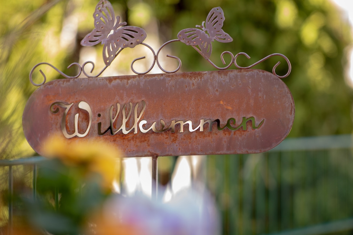 A rustic metal sign displaying the word 'Willkommen' is adorned with decorative butterflies. The sign's weathered texture contrasts against a blurred background of greenery and colorful flowers, inviting guests into the Twin Peaks Lodge.