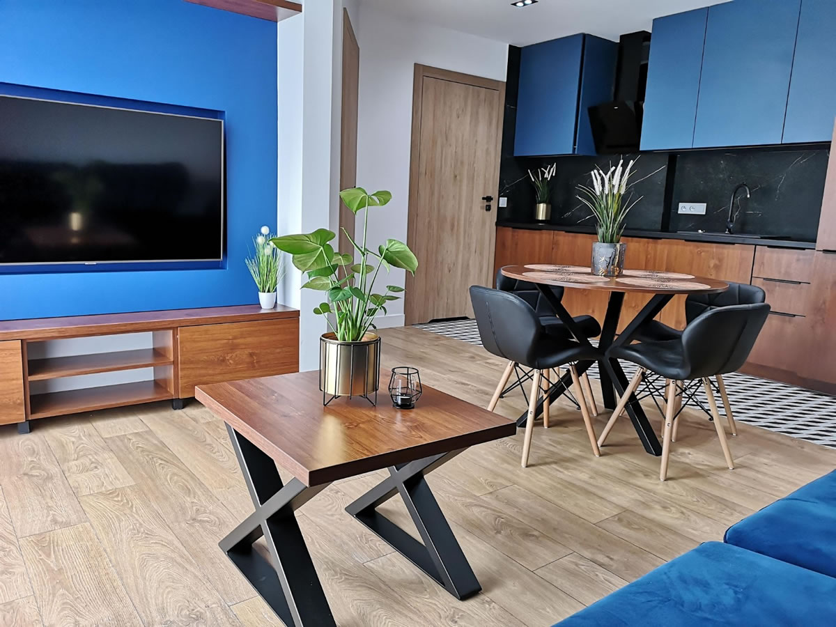 A modern living area features a wooden coffee table with a small plant and candle on it, set against a striking blue accent wall. A dark kitchen area is visible in the background, complemented by a round dining table surrounded by black chairs.