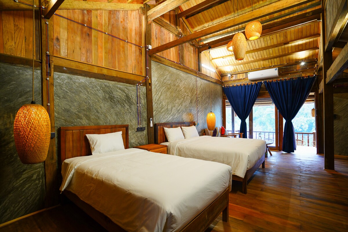 A spacious room features two large beds with crisp white linens. Wooden accents complement the rustic decor, while woven pendant lights provide warm illumination. Dark curtains frame the large window, offering views of the surrounding forest, enhancing the natural ambiance.