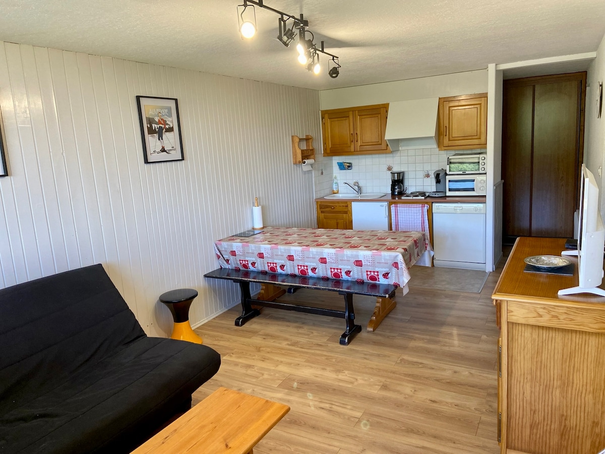 A dining area features a large table with a patterned tablecloth centered in the room. Nearby, a comfortable sofa bed is positioned next to a wooden coffee table. The kitchen is equipped with wooden cabinets and essential appliances, illuminated by track lighting overhead.