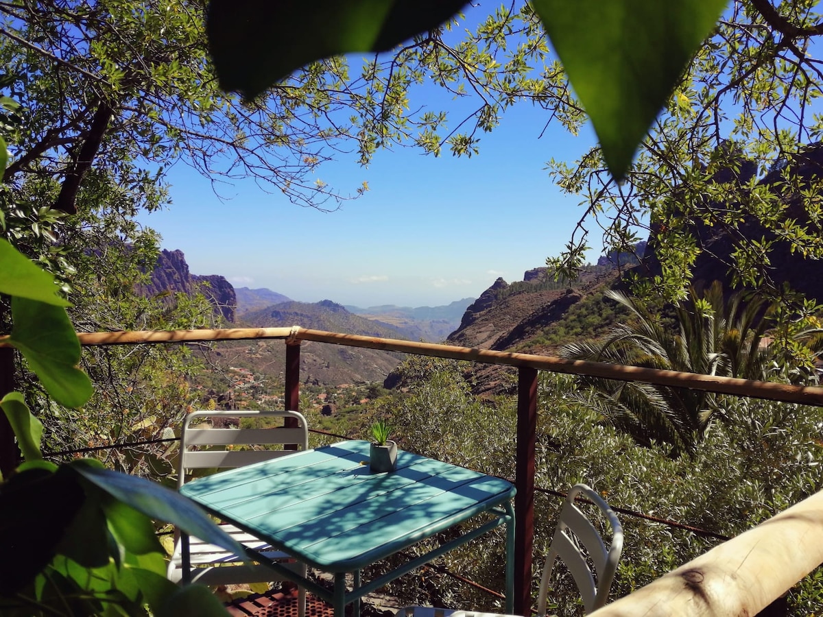 A turquoise table with matching chairs is positioned on a balcony surrounded by lush greenery. A small potted plant rests on the table, and a scenic view of mountains and valleys extends into the horizon, creating a serene outdoor space.