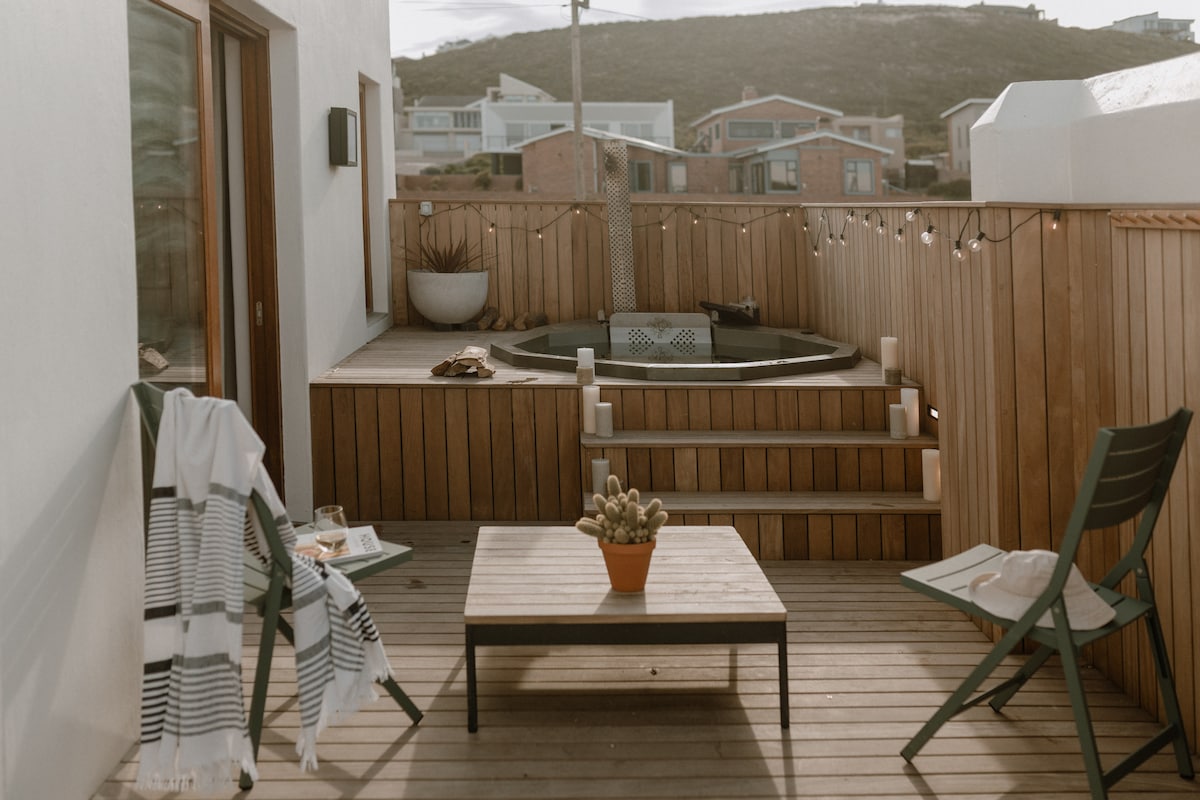 A serene outdoor deck features a hot tub surrounded by wooden decking. Two chairs in a minimalist design face a small black table, adorned with a potted plant. Soft string lights add a gentle ambiance to the inviting space.