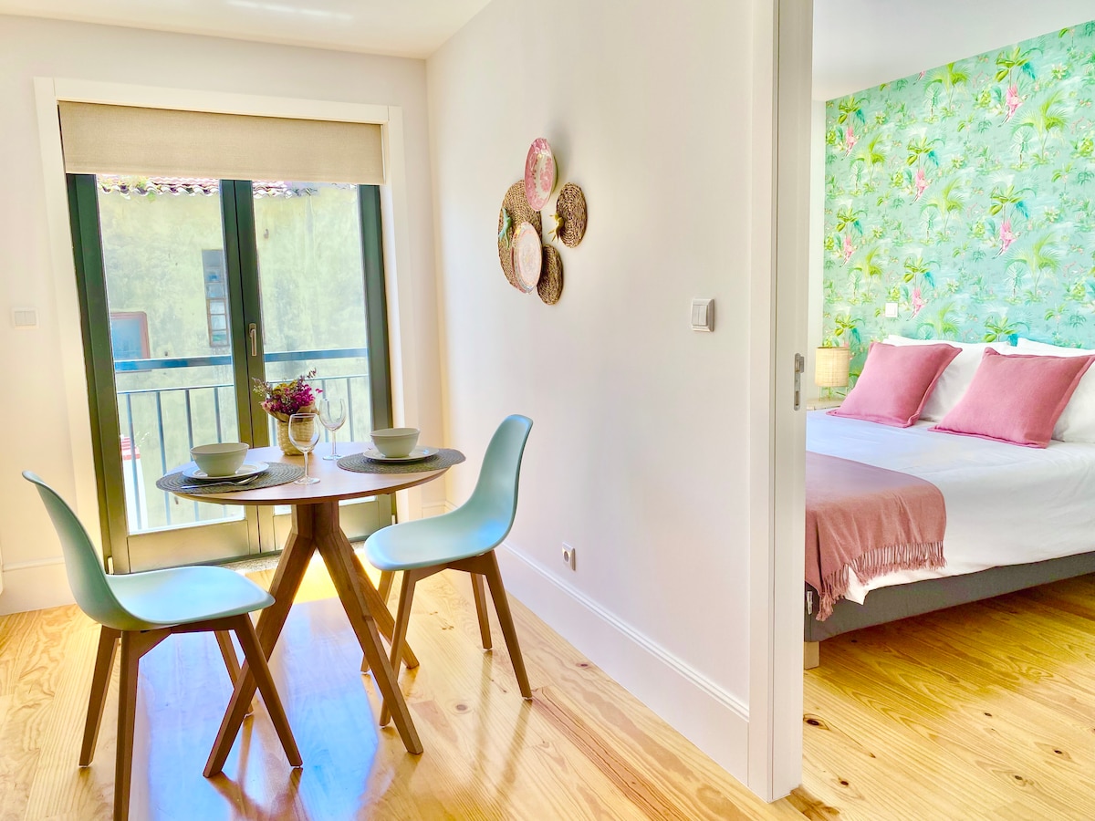 A dining area features a round table with two light blue chairs and table settings for two guests. In the background, a spacious bed with soft pink accents is visible, complemented by a colorful floral wallpaper. Natural light enters through the glass doors leading to a small balcony.