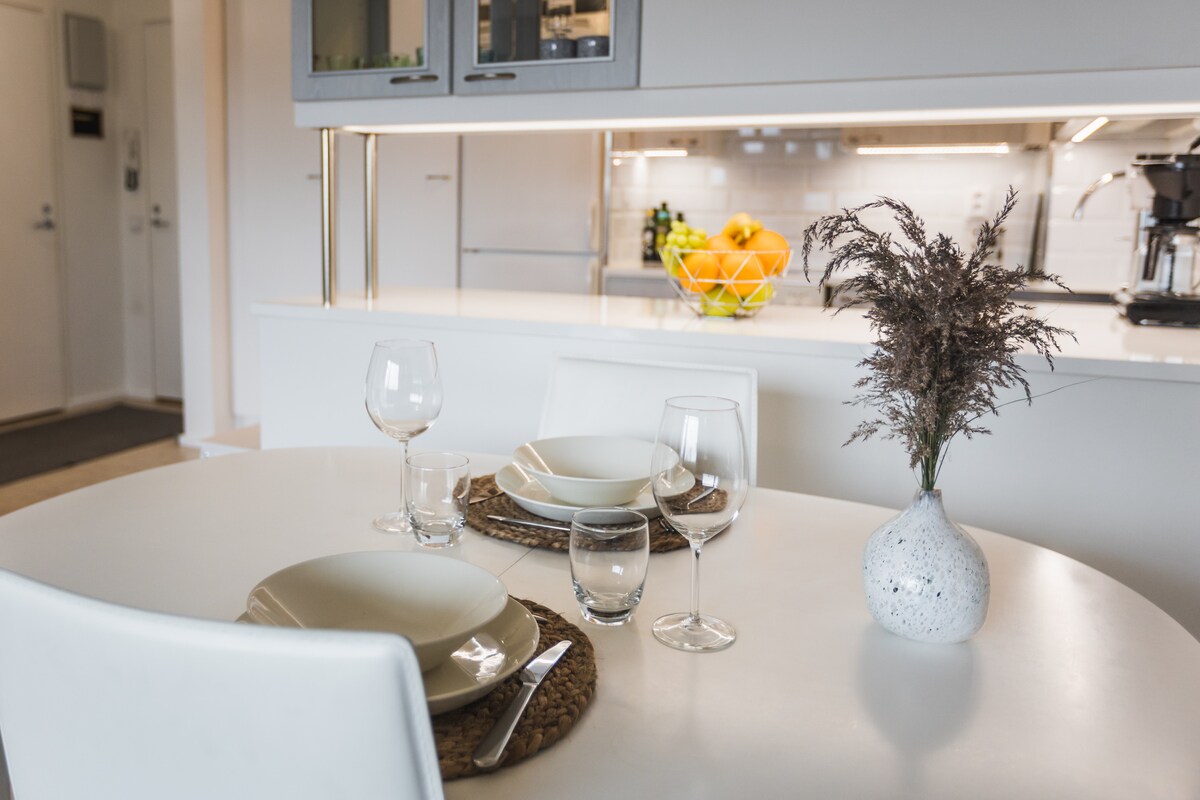 A dining area is presented with a white table set for a meal. Simple plates, glasses, and decorative elements are arranged neatly. In the background, a modern kitchen with bright lighting and a bowl of fresh fruit is visible, enhancing a clean and inviting space.