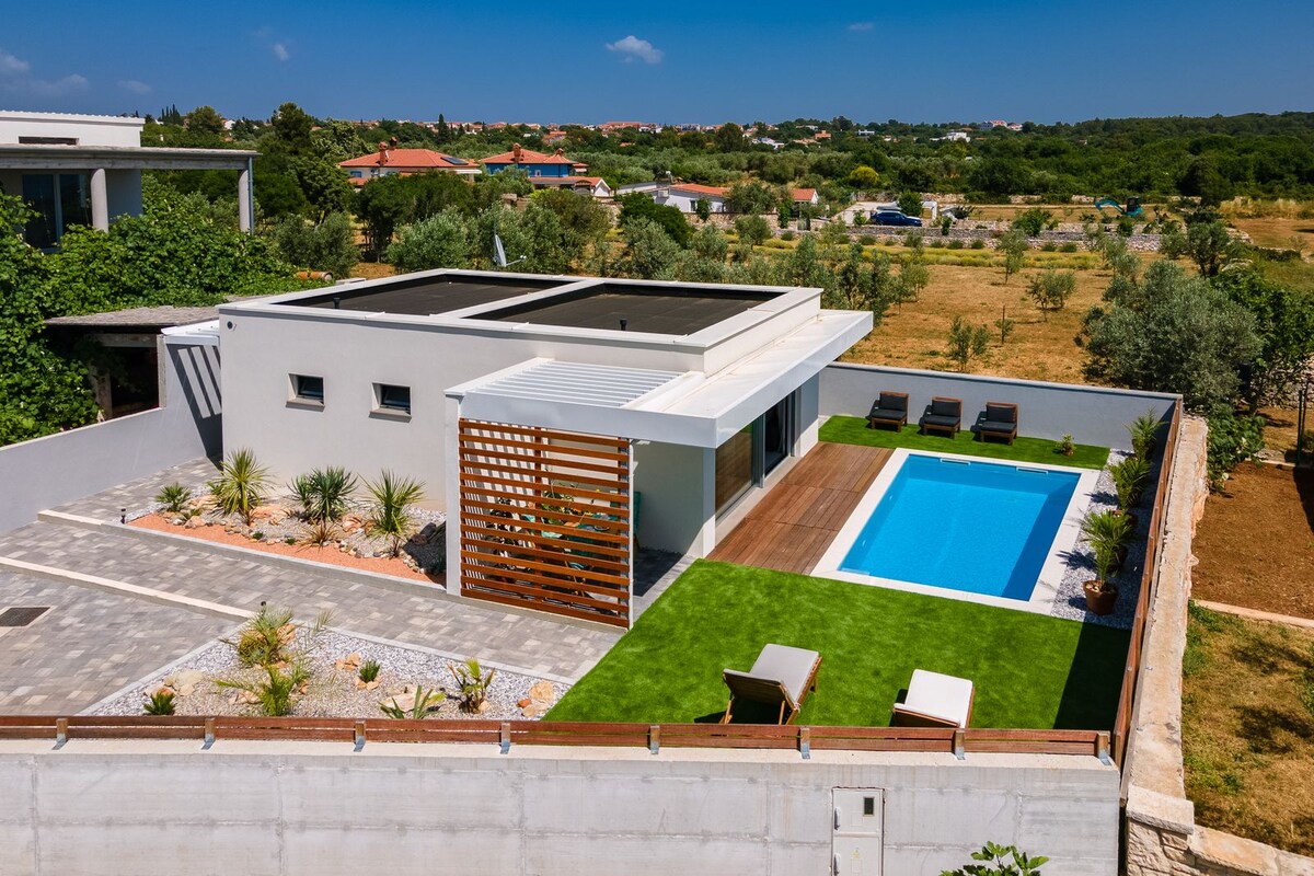 A modern holiday home is shown from an aerial view, featuring a well-maintained yard with a swimming pool surrounded by sun loungers. Lush greenery and landscaped areas complement the structure, while solar panels are visible on the roof, promoting sustainability and comfort.