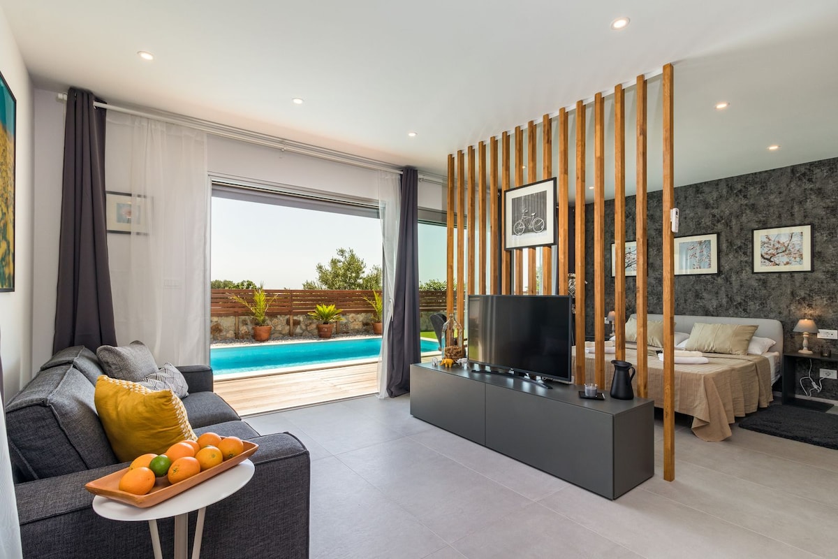 The open living area features a comfortable sofa with decorative cushions, a coffee table with fresh fruit, and a TV on a minimalistic stand. Natural light flows in through large sliding doors, revealing an outdoor area with a swimming pool and greenery.