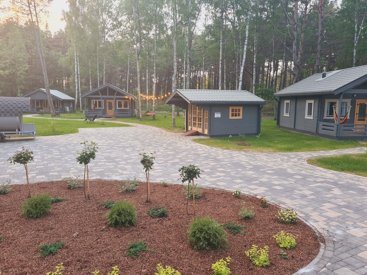 A view of the property features multiple cabins surrounded by greenery. A paved pathway leads through a neatly landscaped area with flower beds and shrubs, providing a serene ambiance. Trees offer natural shade, contributing to the peaceful atmosphere of the setting.