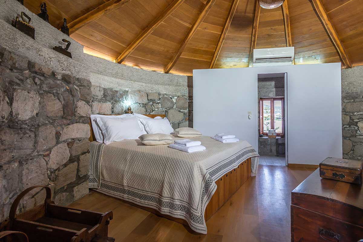 The round bedroom features a spacious bed adorned with neutral blankets and several folded towels. Stone walls provide a rustic charm, complemented by a wooden ceiling. A door leads to the bathroom, while natural light filters in through a nearby window.