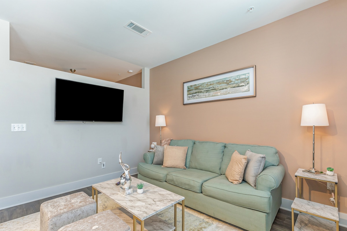 A comfortable living area is presented with a soft green sofa, accentuated by decorative pillows. A mounted flat-screen TV is visible on the wall opposite. Two elegant side tables with minimal decor flank the sofa, while warm lighting is provided by two lamps.