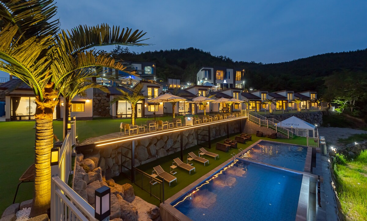 A serene outdoor space is illuminated at dusk, featuring an infinity pool reflecting soft lights. Comfortable lounge chairs line the poolside, with a sloping lawn and palm trees in the background, complemented by the modern architecture of the resort buildings.