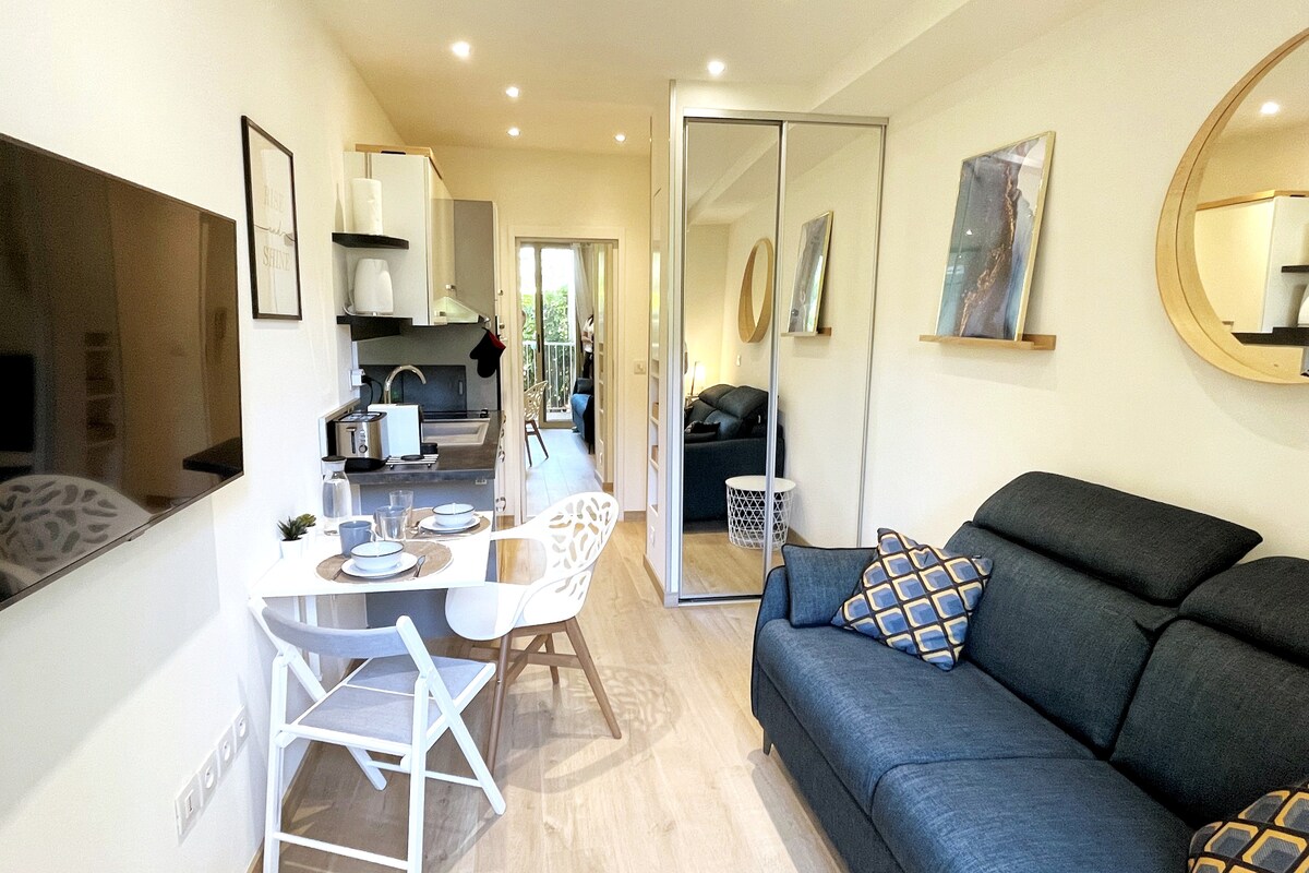 A modern studio is depicted featuring a comfortable sofa and a round dining table set for two. Natural light fills the space through glass doors that lead to a private terrace. The kitchenette is visible, along with tasteful decor that complements the open layout.