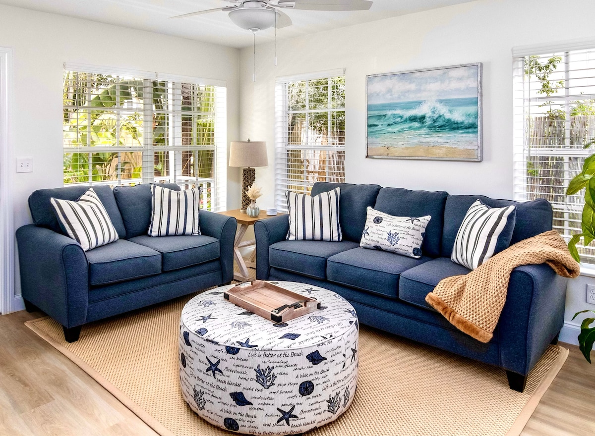 A light-filled living area is furnished with a navy blue sofa set featuring striped pillows. A round coffee table adorned with nautical-themed fabric complements the decor. Natural light streams in through large windows, highlighting a coastal artwork on the wall.