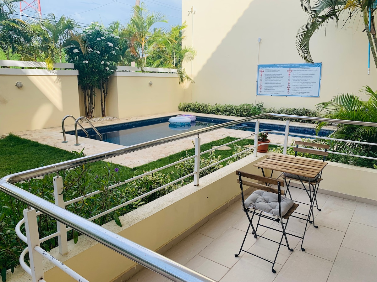 A well-maintained outdoor area is visible, featuring a small pool surrounded by greenery. A two-person table made of wood is positioned nearby, with padded chairs adding comfort. The space is framed by lush plants, creating a relaxing environment for residents.